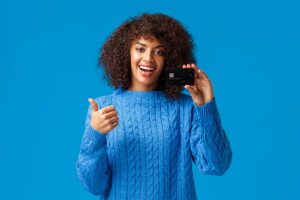 A smiling young African woman wearing a bright blue sweater holds up a black credit card and gives a thumbs-up, symbolizing confidence and empowerment through access to credit cards.