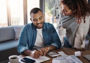 A smiling man takes notes while managing household finances with his partner, surrounded by bills, a calculator, and coffee mugs—illustrating how microcredit empowers families to plan and build a stable financial future together.