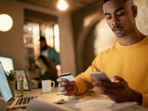 A young man sits at a desk reviewing his finances with a credit card in one hand and a smartphone in the other, symbolizing the importance of conscious credit management and responsible financial decision-making in the digital age.