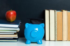 A blue piggy bank wearing a graduation cap sits between stacks of books and notebooks, symbolizing the importance of financial education for students in academic settings.