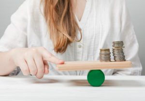 A person balancing stacks of coins on a wooden seesaw, symbolising the careful evaluation of interest rates and financial decisions to achieve stability and fairness.