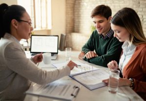 O ChatGPT disse: A financial advisor meeting with a young couple, reviewing documents and explaining options for Loans with Movable Collateral, with a laptop and architectural plans visible on the table.