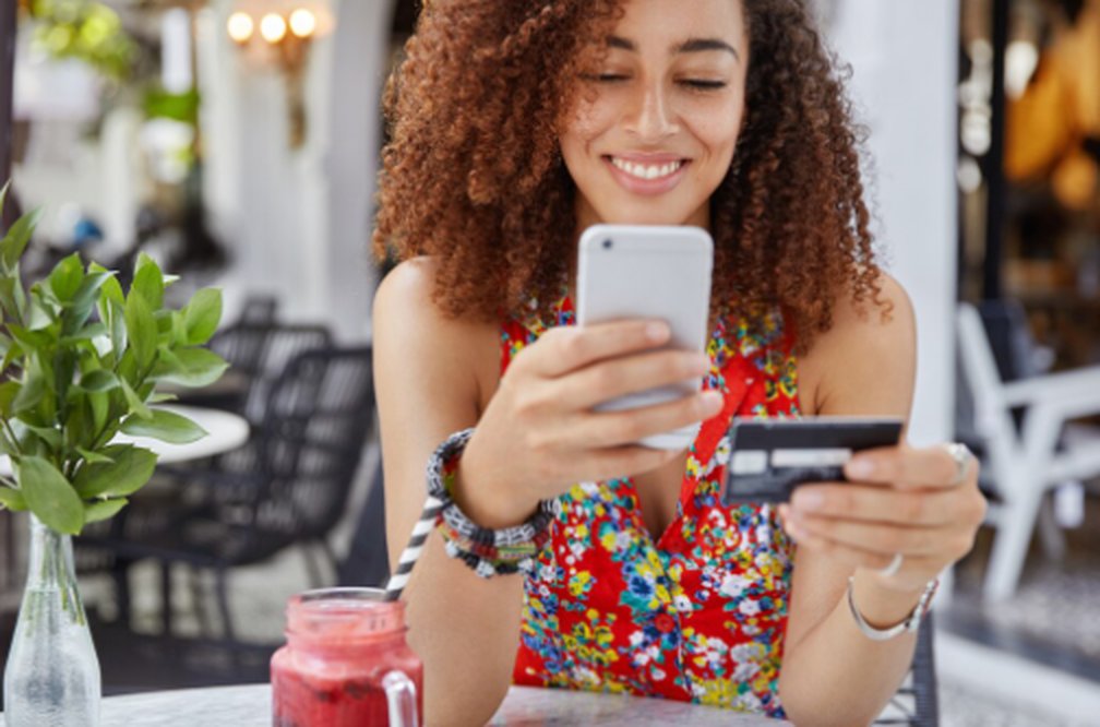 A young woman sits at an outdoor café, smiling as she uses her smartphone and bank card, illustrating the convenience and accessibility of Mobile Money for modern personal finance management.