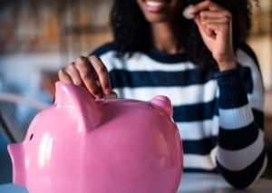 Woman smiling while placing coins into a pink piggy bank, symbolizing smart savings habits and the importance of building an emergency fund for financial security.