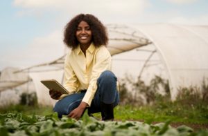 Smiling female farmer in a yellow shirt holding a tablet while inspecting crops in front of a greenhouse, representing the benefits of rural loans for agricultural development.