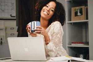 Smiling African woman working on a laptop at home office, holding a coffee mug, representing confidence and progress toward female economic independence.
