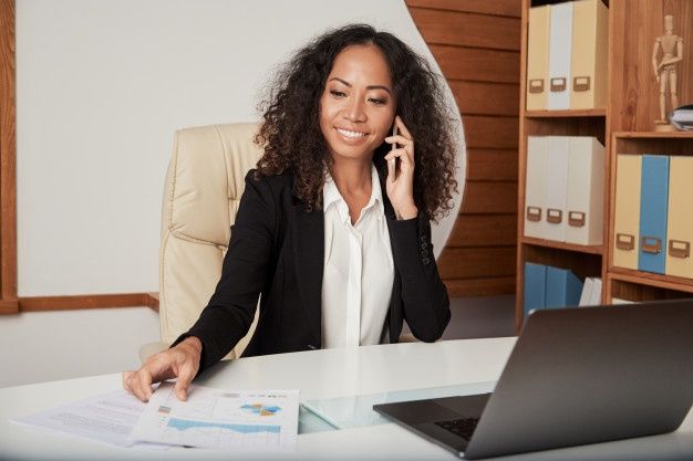 Businesswoman smiling while talking on the phone in her office, analyzing financial charts and working on her laptop, representing success in small businesses.