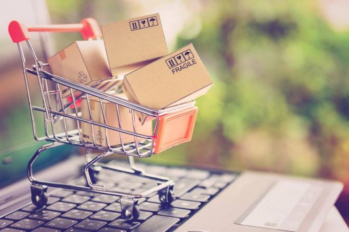 A miniature shopping cart filled with small cardboard boxes labeled “fragile” sits on a laptop keyboard, symbolizing online shopping and the impact of global inflation on consumer purchasing power.